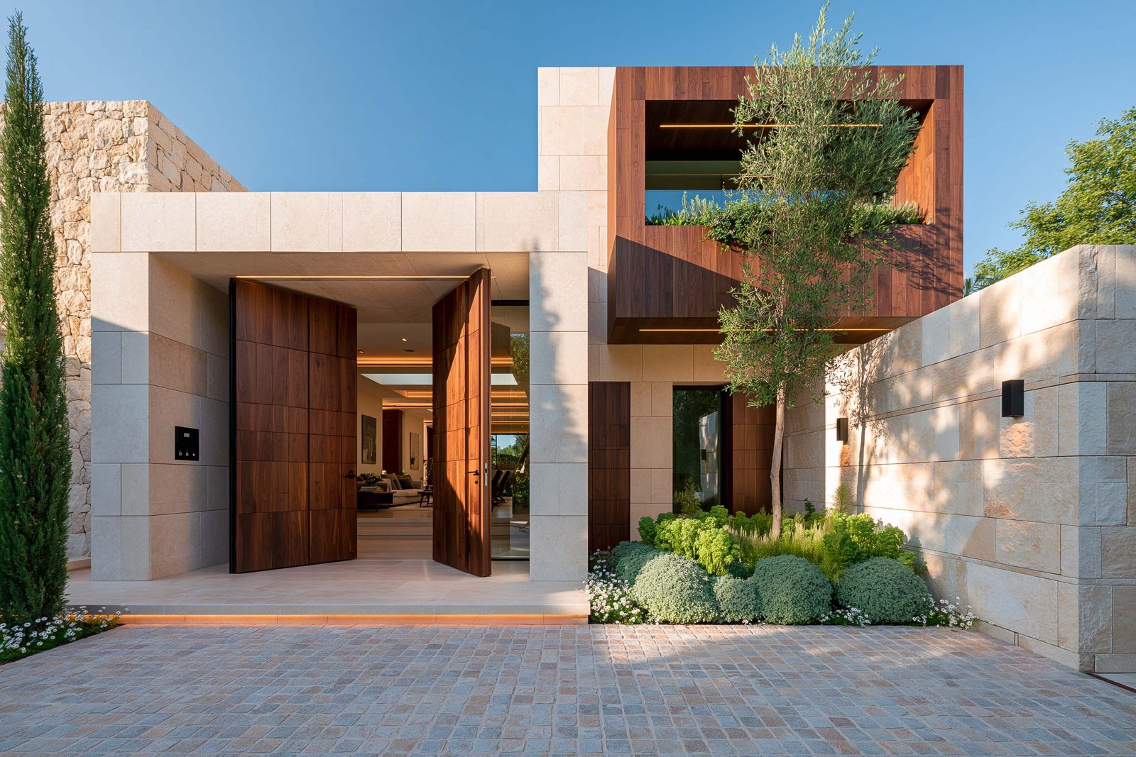 Profondeur et calme — seuil extérieur d’une villa contemporaine à Tourrettes-sur-Loup en plein midi, façade en pierre claire calcaire et double porte pivotante en noyer fumé, Cupressus sempervirens stricta en port colonne, Phlomis fruticosa et Santolina chamaecyparissus en pied de mur avec Erigeron karvinskianus, ciel bleu profond, Paracelsus Gardens™