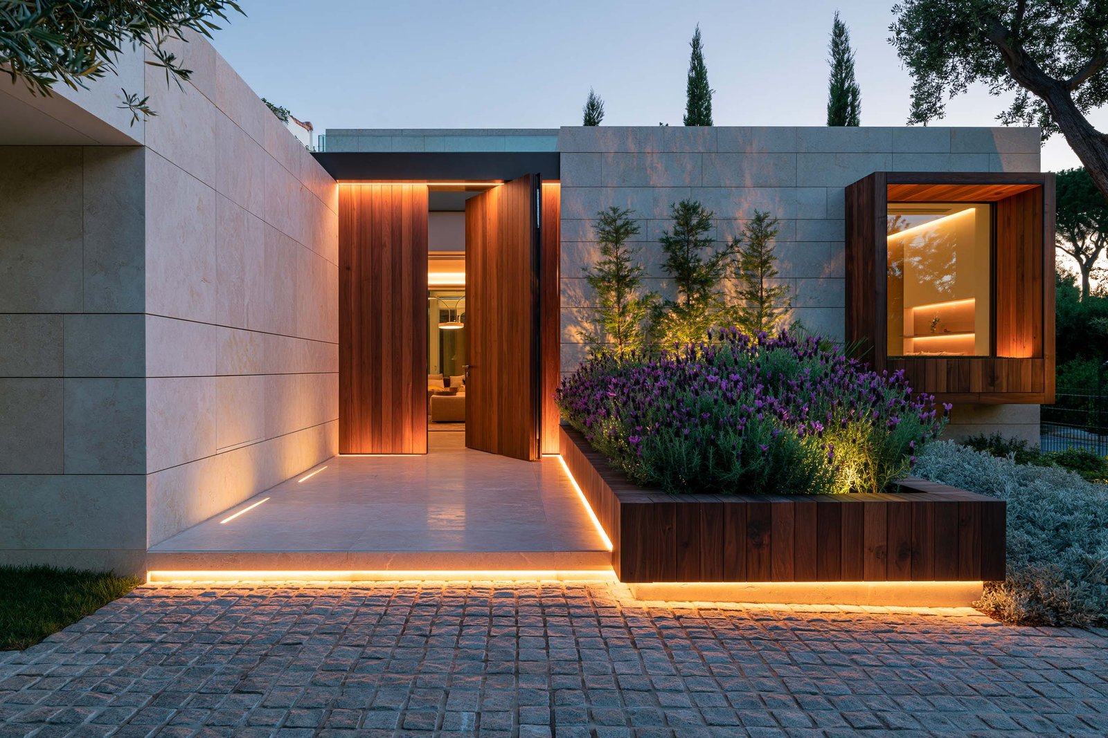 Seuil — entrée et cadrage : seuil extérieur d’une villa au Cap-Ferrat au crépuscule, porte pivotante en noyer fumé, jardinière Juliet™ Riviera Classica en noyer fumé portant Lavandula stoechas en floraison violette, Cupressus sempervirens stricta et Olea europaea visibles au-delà, pavés cobblestone gris, LED 2700K cachée, Paracelsus Gardens™