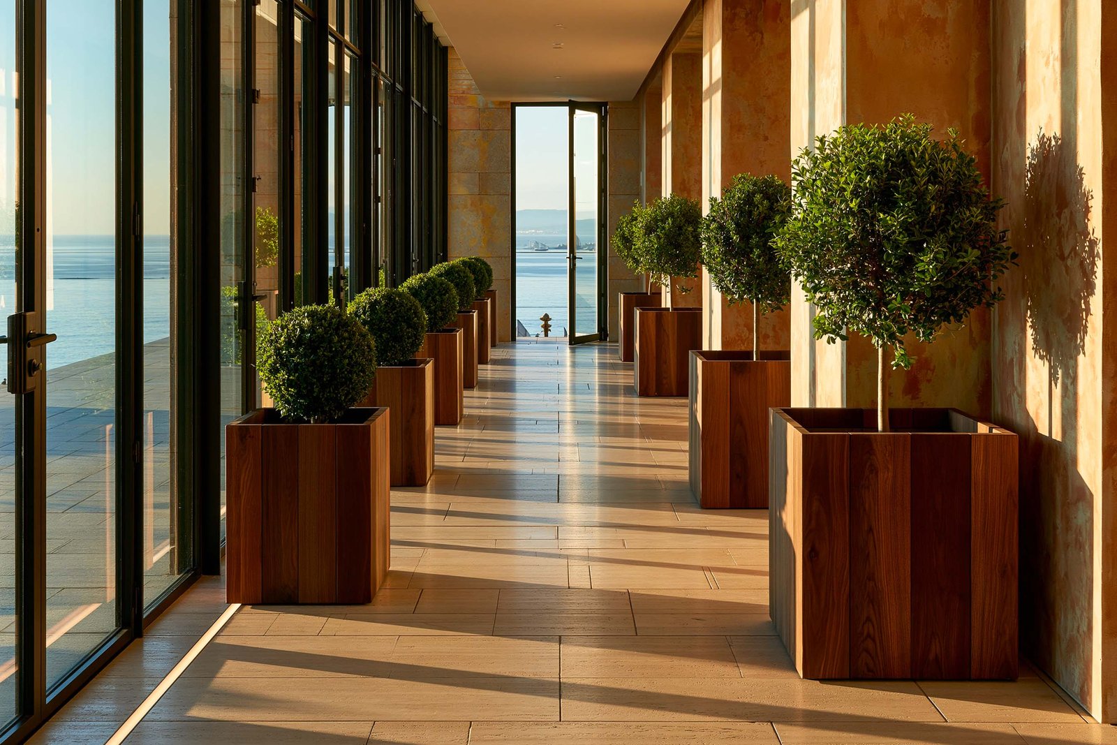 Guidance par le rythme — longue enfilade d’un hôtel boutique à Monaco au golden hour, suite régulière de planters Lignum™ Block en noyer fumé alignés sur sol travertin clair guidant l’œil vers une porte vitrée ouverte sur l’horizon Méditerranée, mur stucco lustro ocre patiné à droite et baie vitrée à gauche, Buxus sempervirens en topiaires boules dans les planters, LED 2700K cachée linéaire au sol, ombres portées rythmant la perspective, Paracelsus Gardens™