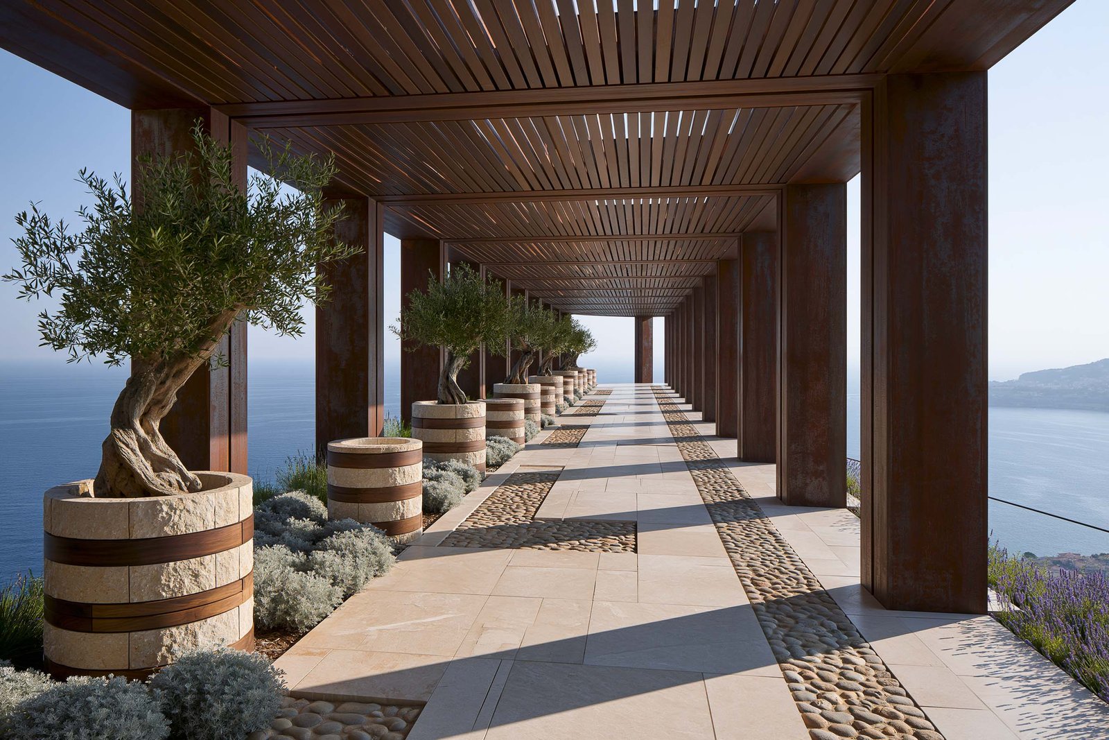 Roquebrune-Cap-Martin — pergola d’une villa de bord de mer, colonnade rythmique en acier patiné à chaud sous plafond lattes en bois, alignement de Lignum™ Petra cylindriques en pierre claire cerclée de bandes de noyer fumé accueillant Olea europaea anciens sculpturaux, Lavandula angustifolia en masses basses et Helichrysum italicum argenté, dalle en pierre claire et calade ouverte sur la Mer Méditerranée — Paracelsus Gardens™