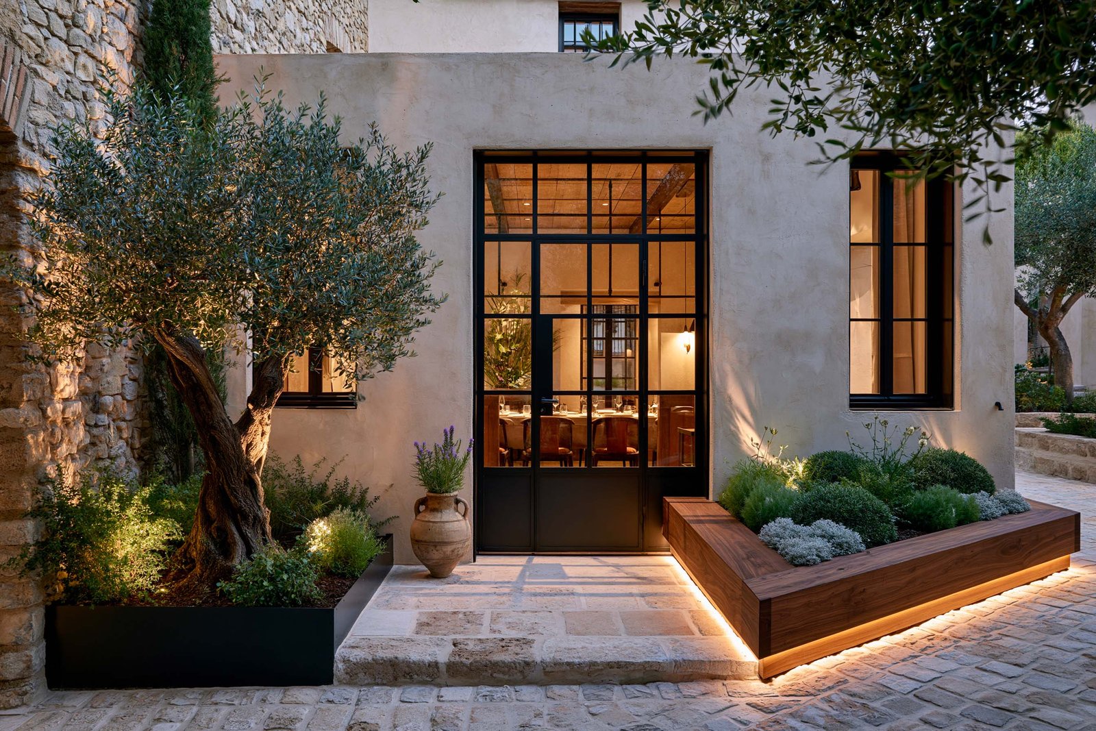 Seuil extérieur d’un restaurant à Saint-Paul-de-Vence, porte rectangulaire en acier noirci dans mur chaulé, olivier à gauche et jardinière Lignum en noyer fumé plantée de lavande et helichrysum à droite.