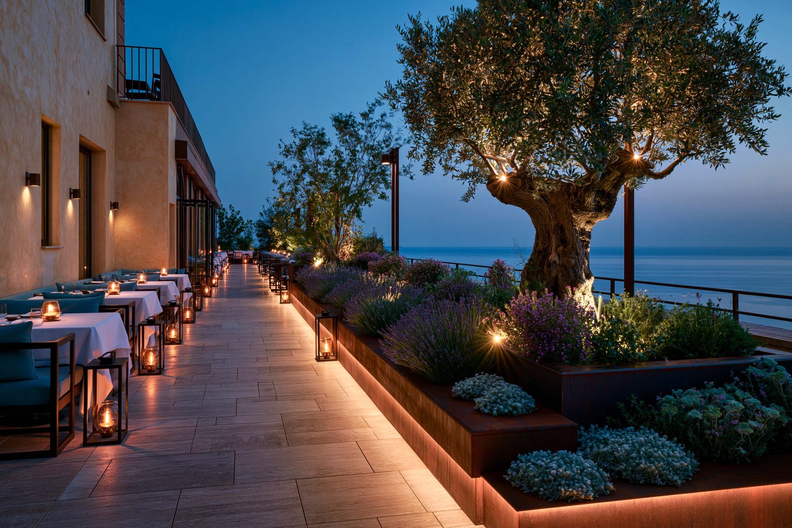 Terrasse de restaurant à Roquebrune-Cap-Martin à l’heure bleue, séquence de tables nappées blanc aux bougies, jardinières basses plantées de lavande et helichrysum, olivier mature éclairé en LED chaude.