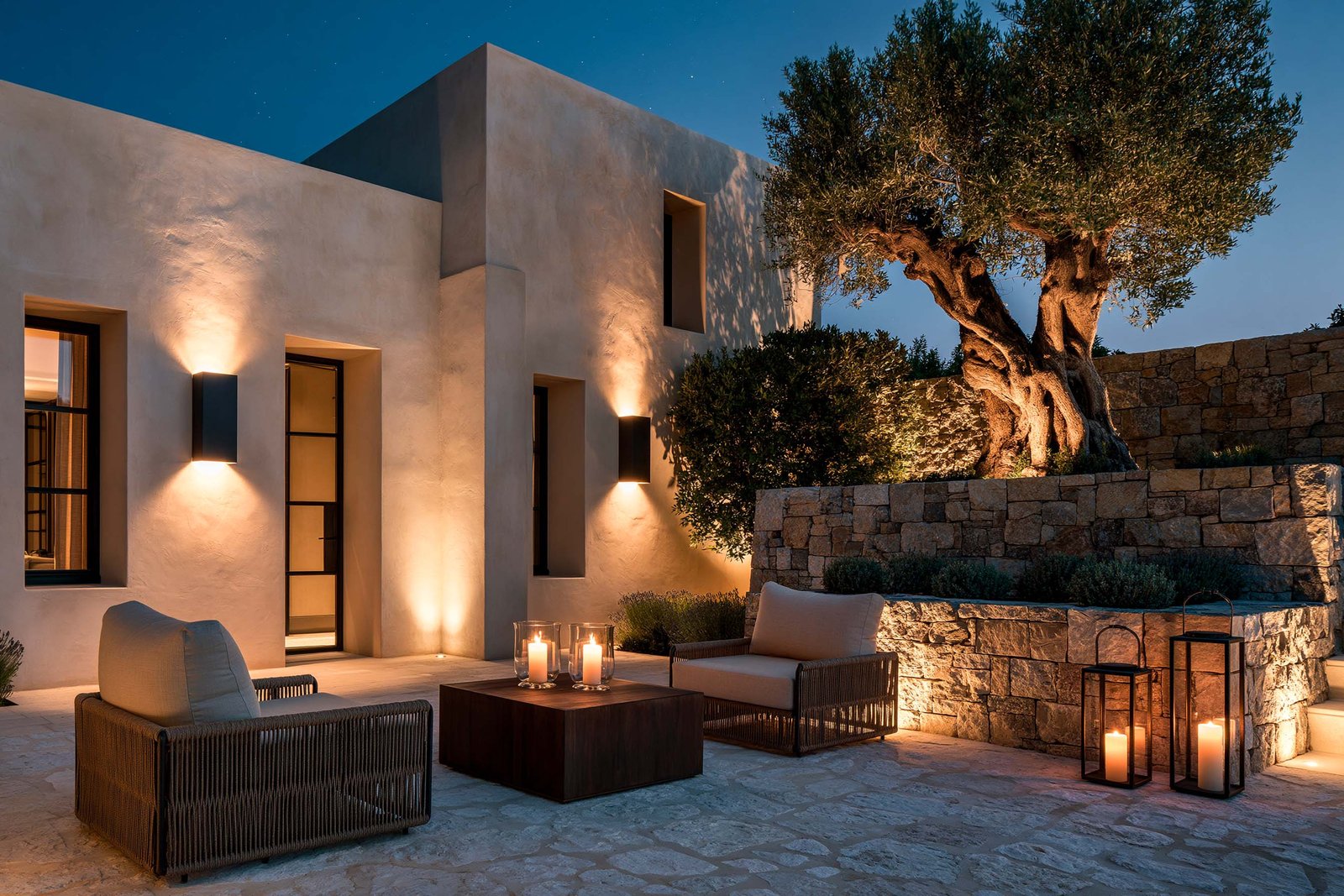 Patio nocturne d’une villa à Saint-Paul-de-Vence, fauteuils outdoor bas en lin écru sur une cour pavée en pierre claire, table basse en noyer fumé portant des bougies en verre soufflé, façade en stucco lustro chaud rétro-éclairée, lanternes au sol, ciel étoilé.