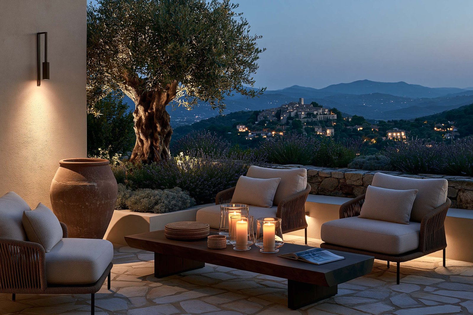Salon de terrasse au crépuscule d’une villa à Èze, deux fauteuils outdoor bas en lin écru sur acier noirci, table basse en noyer fumé avec bougies en verre soufflé, masses de Lavandula angustifolia, vue sur un village perché de l’arrière-pays méditerranéen à la blue hour.